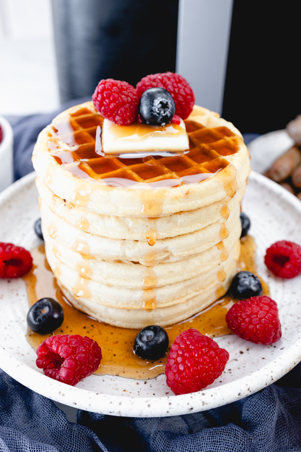 Close up view of a stack of waffles on a plate, topped with syrup, butter, and berries. Behind the plate is an air fryer.
