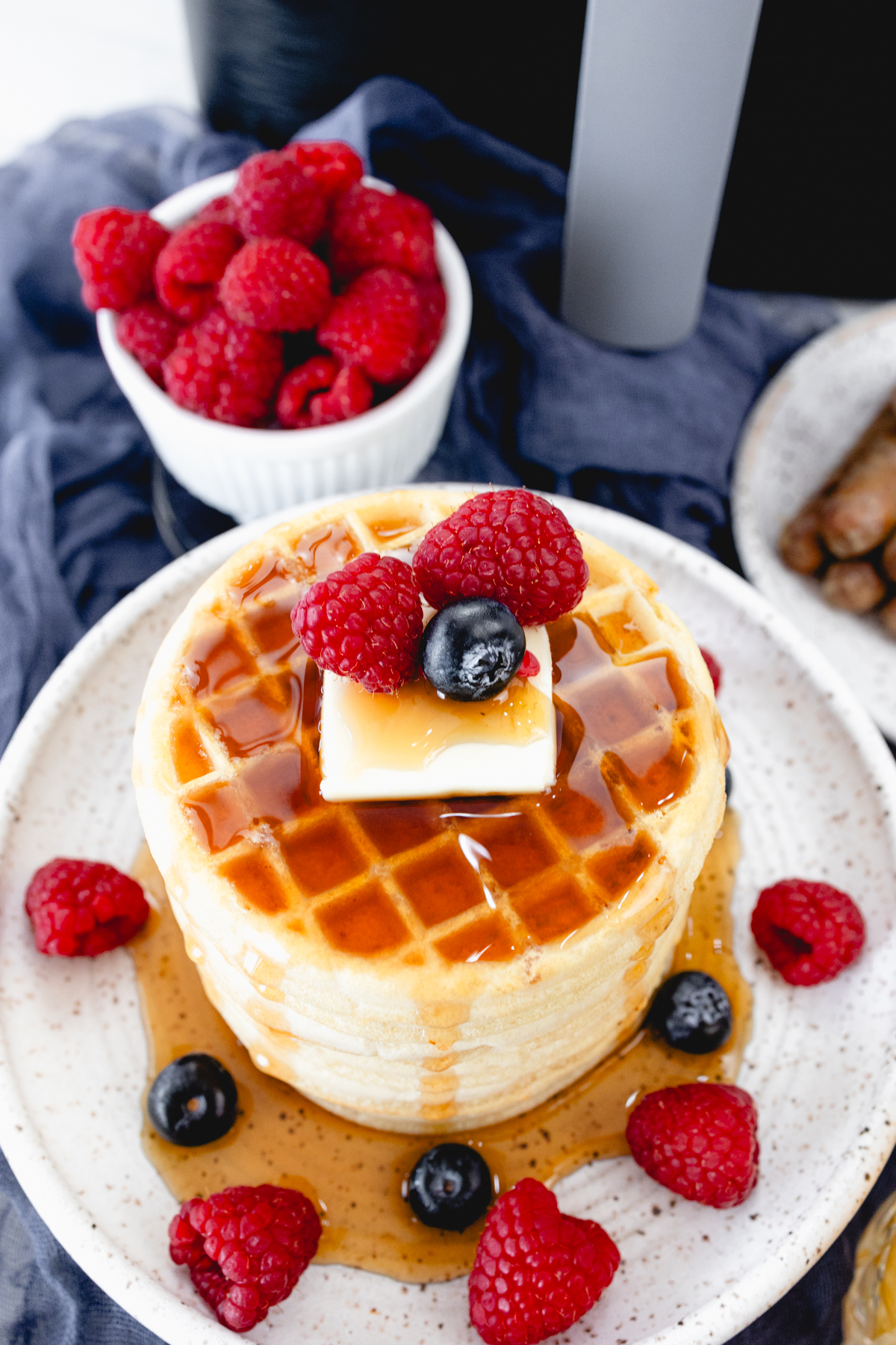 Close up view of a stack of waffles on a plate, topped with syrup, butter, and berries. Behind the plate is an air fryer and a bowl of berries.