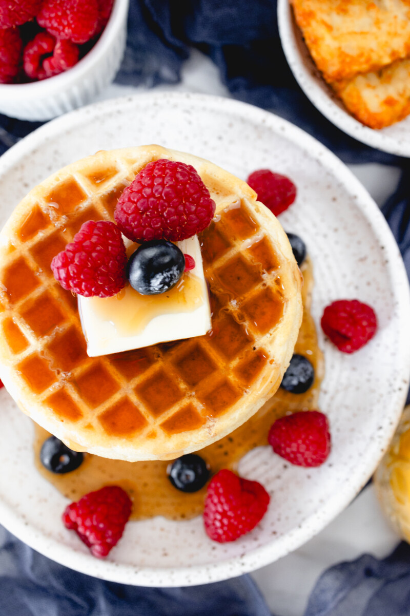 Top view of a stack of waffles on a plate, topped with syrup, butter, and berries. Behind the plate is an air fryer, a bowl of berries, and a plate of hash browns.