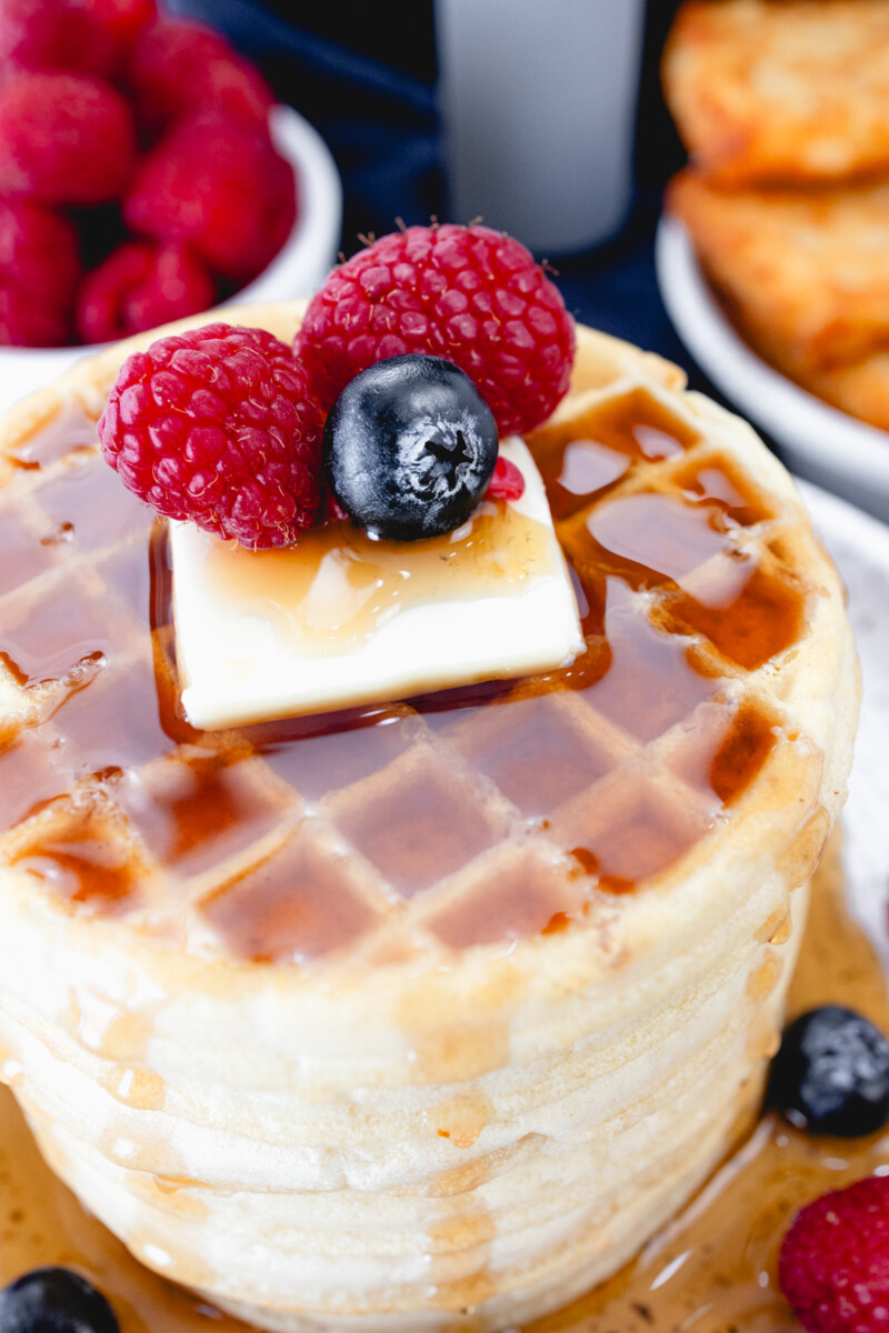Close up view of a stack of waffles on a plate, topped with syrup, butter, and berries. Behind the plate is an air fryer, a bowl of berries, and a plate of hash browns.