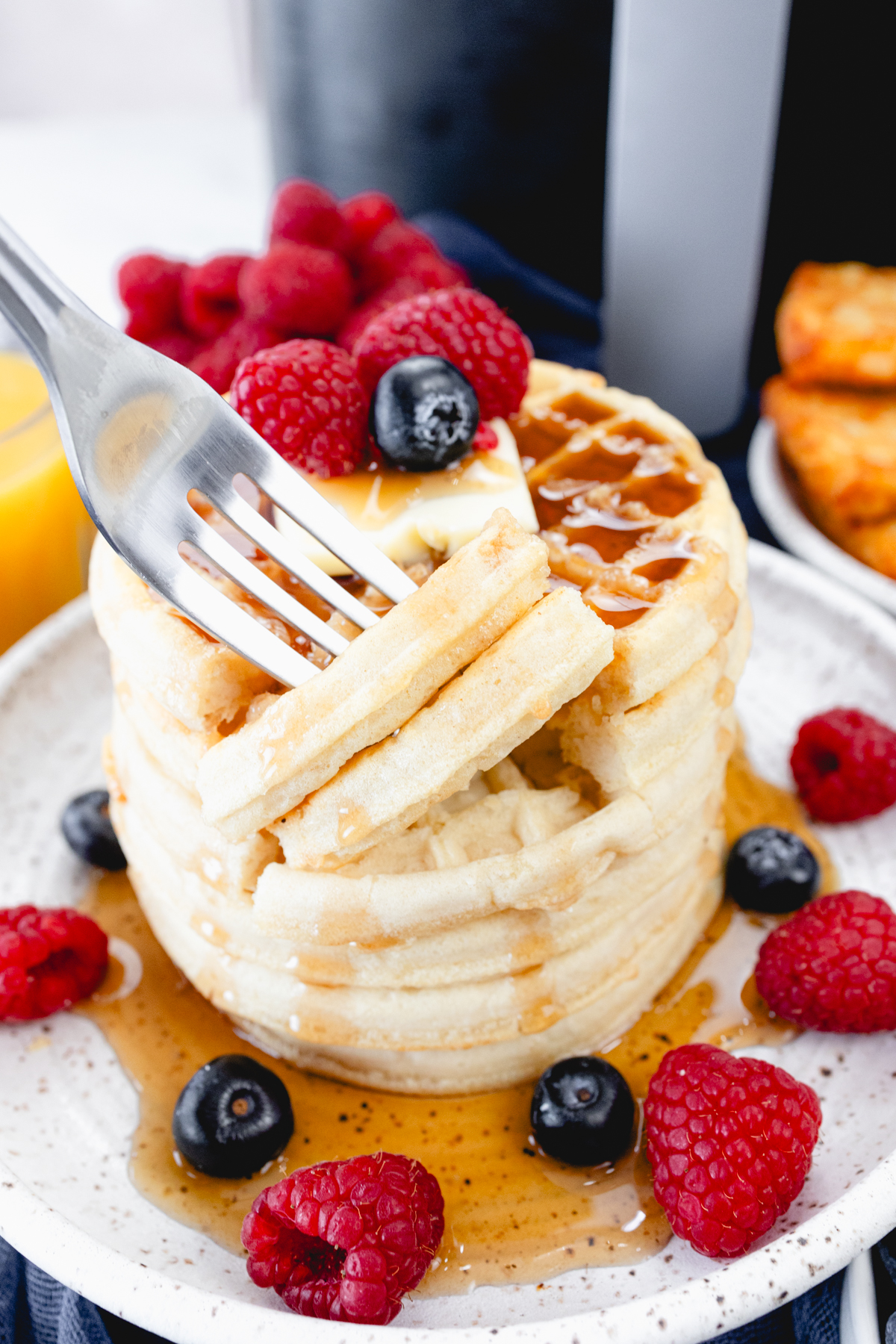 Close up view of a stack of waffles on a plate, topped with syrup, butter, and berries. A fork is in the foreground with skewered pieces of waffle on it. Behind the plate is an air fryer, a bowl of berries, a glass of orange juice, and a plate of hash browns.