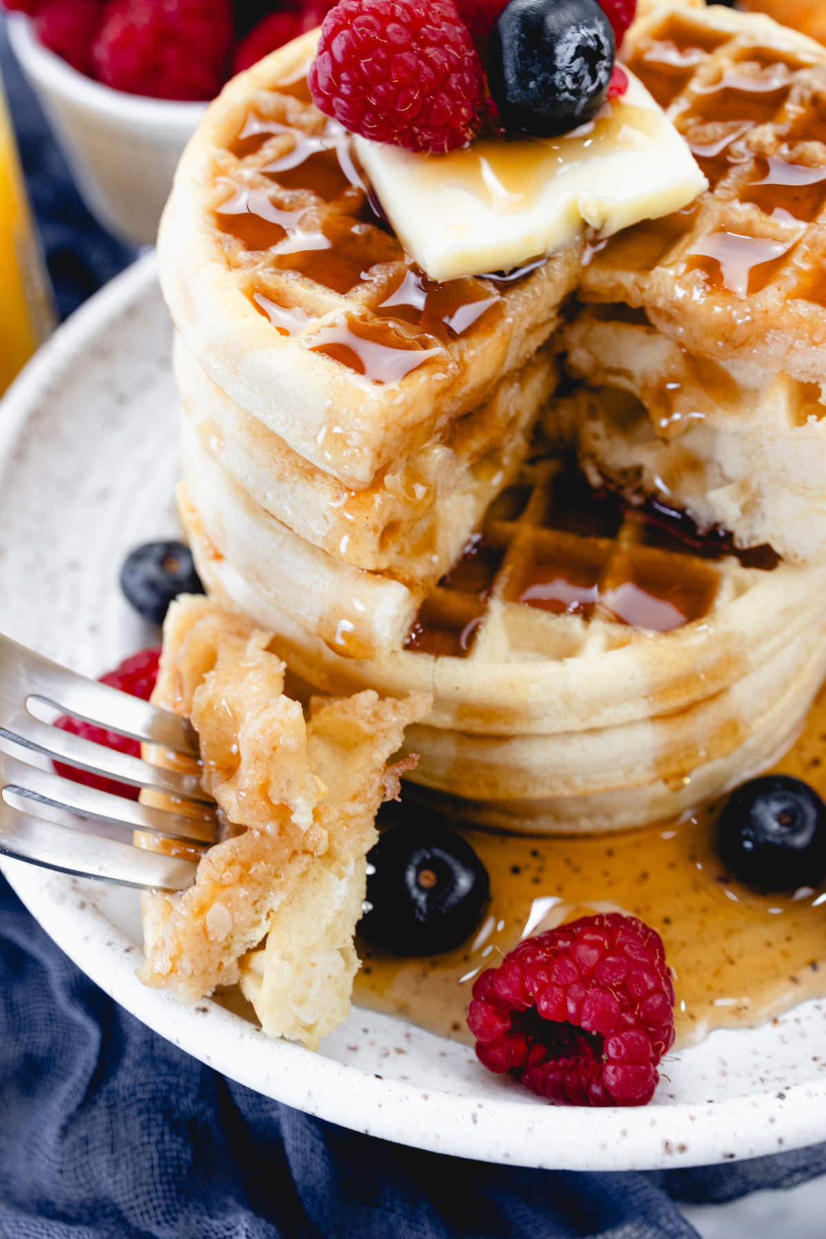 Close up view of a stack of waffles on a plate, topped with syrup, butter, and berries. A fork is in the foreground with skewered pieces of waffle on it. Behind the plate is a bowl of berries, and a glass of orange juice.