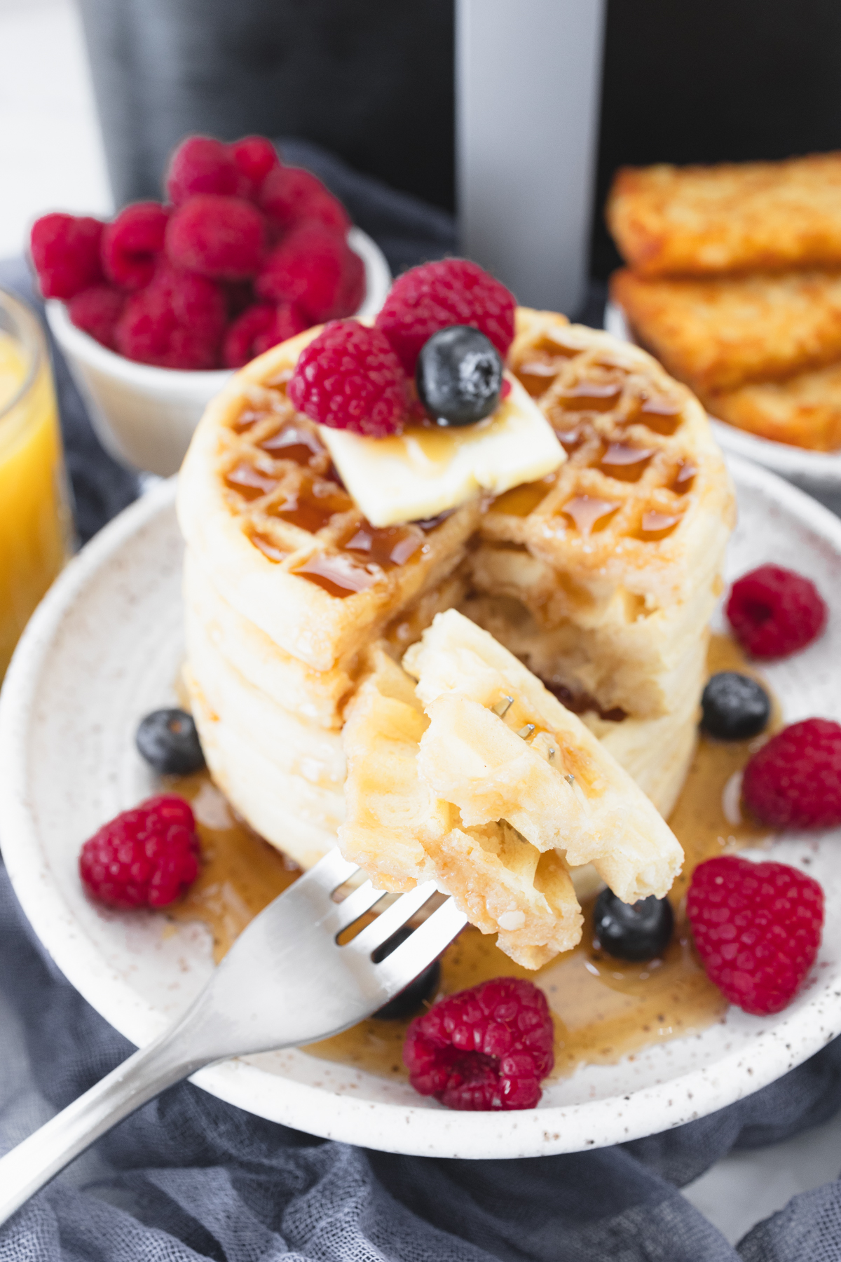 Close up view of a stack of waffles on a plate, topped with syrup, butter, and berries. A fork is in the foreground with skewered pieces of waffle on it. Behind the plate is an air fryer, a bowl of berries, a glass of orange juice, and a plate of hash browns.