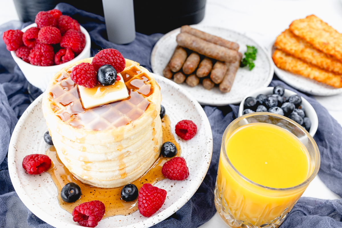 An image of a full breakfast spread featuring: a stack of waffles on a plate in the foreground, topped with syrup, butter, and berries; a glass of orange juice; a bowl of berries; a plate of sausages; and a plate of hash browns. There is an air fryer in the background.