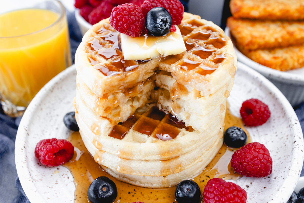 Close up view of a stack of waffles on a plate, topped with syrup, butter, and berries. Half of the waffles have had a triangular quarter cut out of them. Behind the plate is an air fryer, a bowl of berries, a glass of orange juice, and a plate of hash browns.