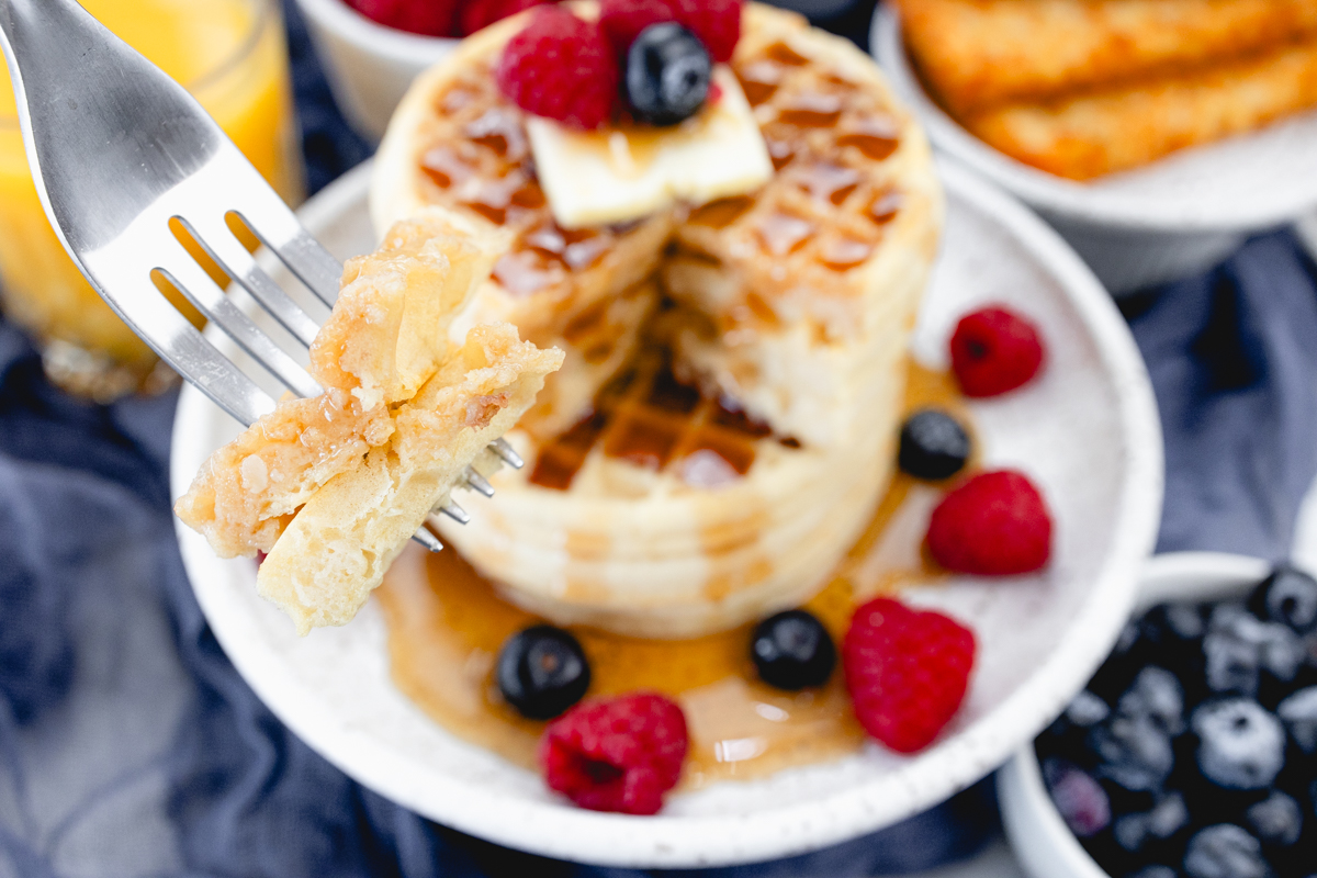 Close up view of a stack of waffles on a plate, topped with syrup, butter, and berries. A fork is in the foreground with skewered pieces of waffle on it. Behind the plate is an air fryer, a bowl of berries, a glass of orange juice, and a plate of hash browns.