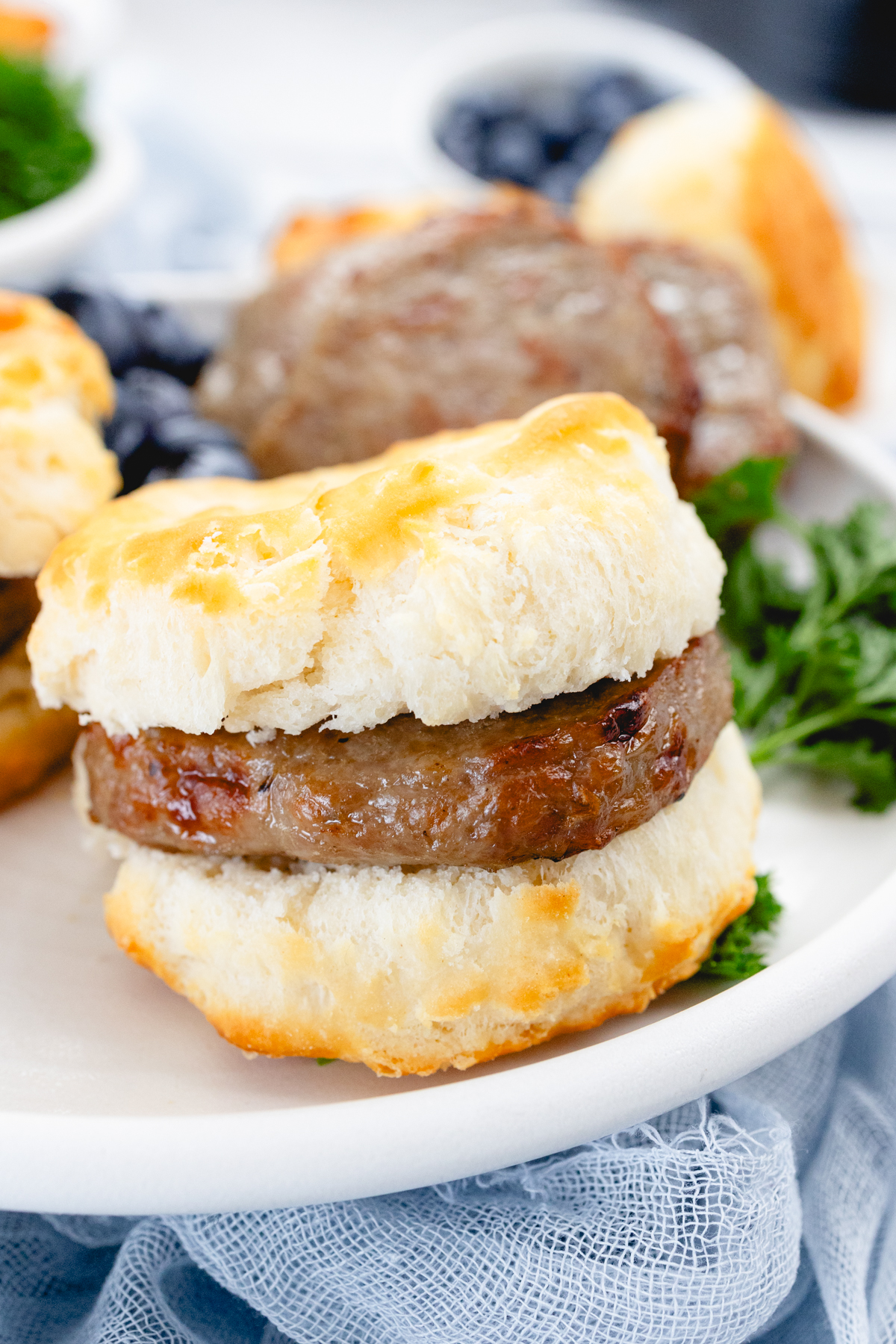 Close up of cooked sausage patties as breakfast sandwiches in sliced biscuits, on a white plate with a leafy green garnish and blueberries on the side.