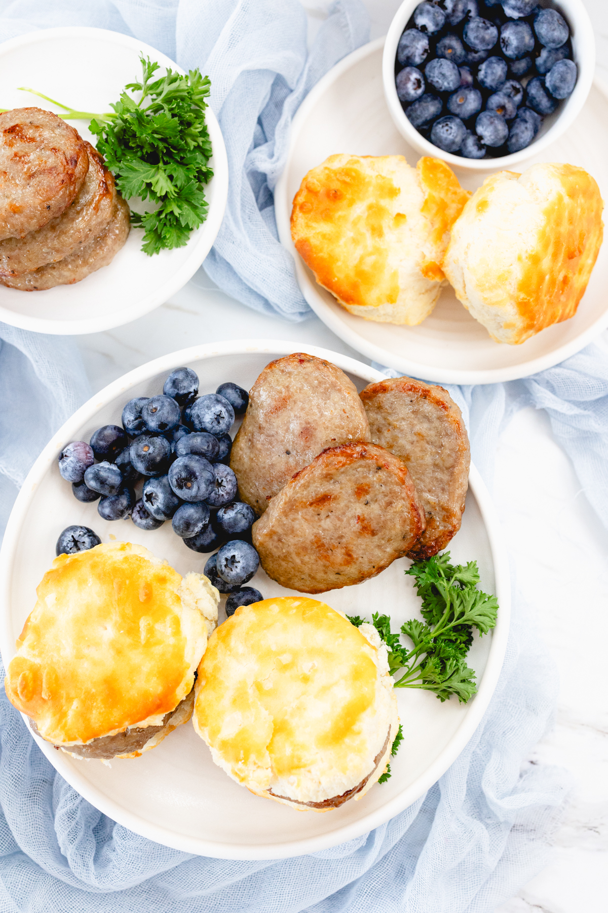 Top view of cooked sausage patties on a white plate with a leafy green garnish, blueberries, and biscuits on the side.