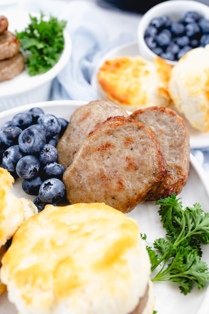 Close up of cooked sausage patties on a white plate with a leafy green garnish, blueberries, and biscuits on the side.