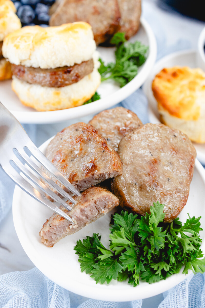 Top view of cooked sausage patties on a white plate with a leafy green garnish to the side, and a fork has skewered a sliced half of one of the patties.