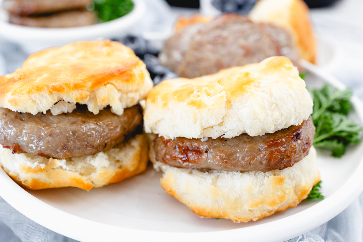 Close up of cooked sausage patties as breakfast sandwiches in sliced biscuits, on a white plate with a leafy green garnish and blueberries on the side.