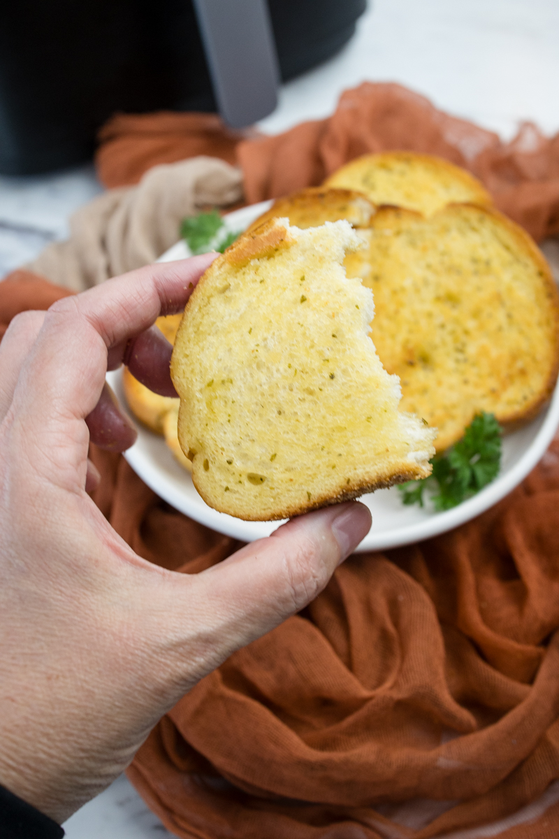Close up view of a hand holding up half a slice of Texas Toast in front of more Texas Toast slices stacked on a white plate.