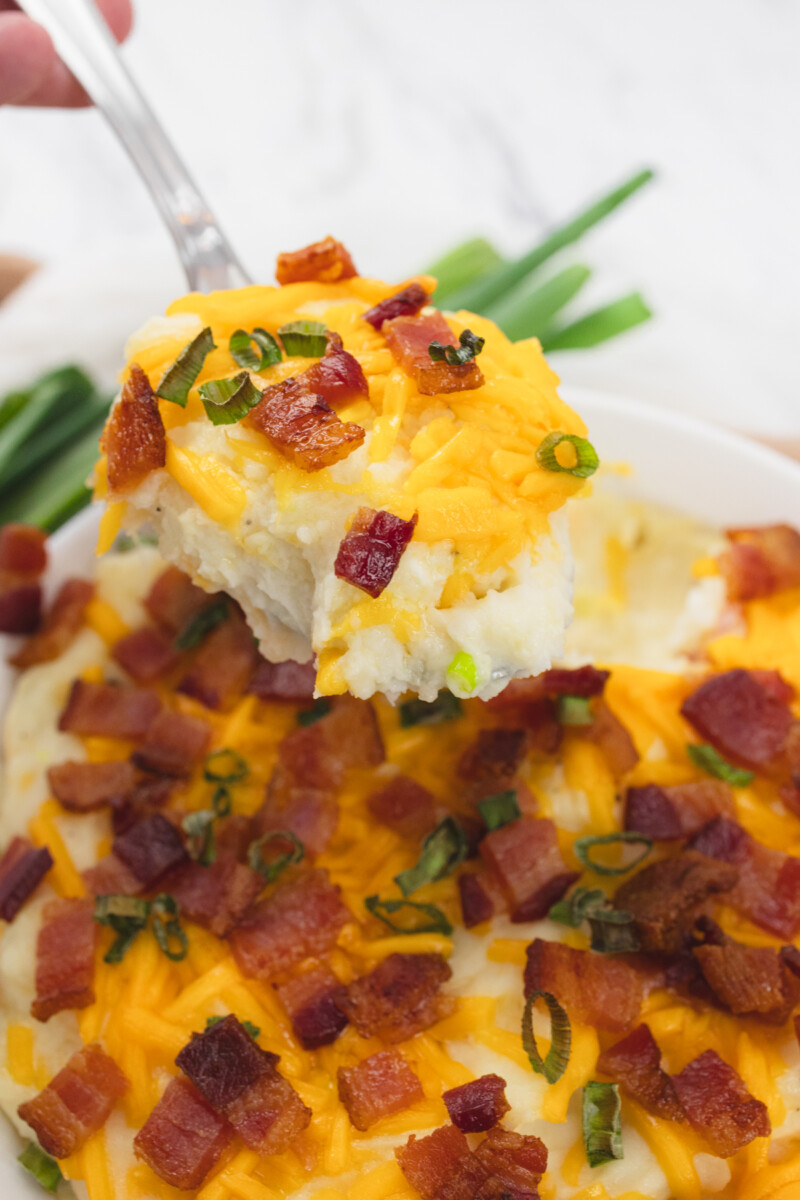 Close up view of a serving spoon lifting a portion of loaded mashed potatoes out of the dish.