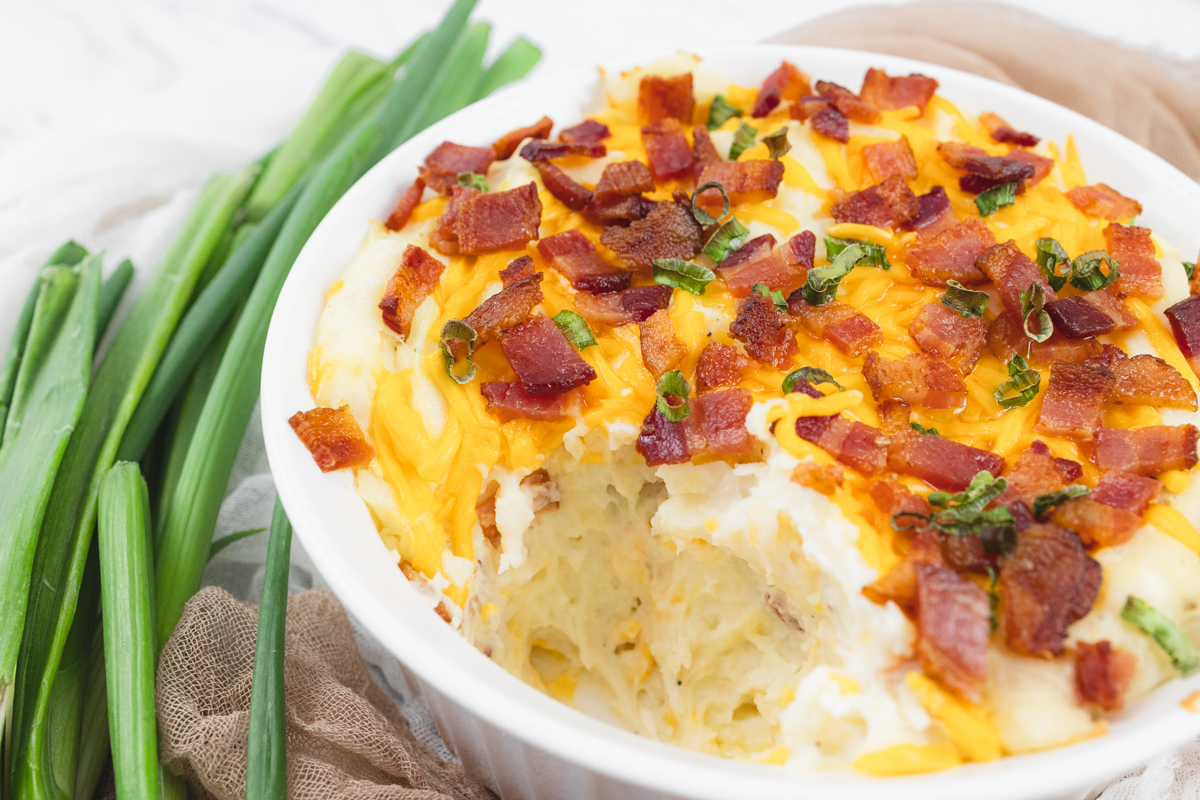 Close up view of a white dish filled with loaded mashed potatoes where a portion has been removed to show the topping and the potatoes underneath.