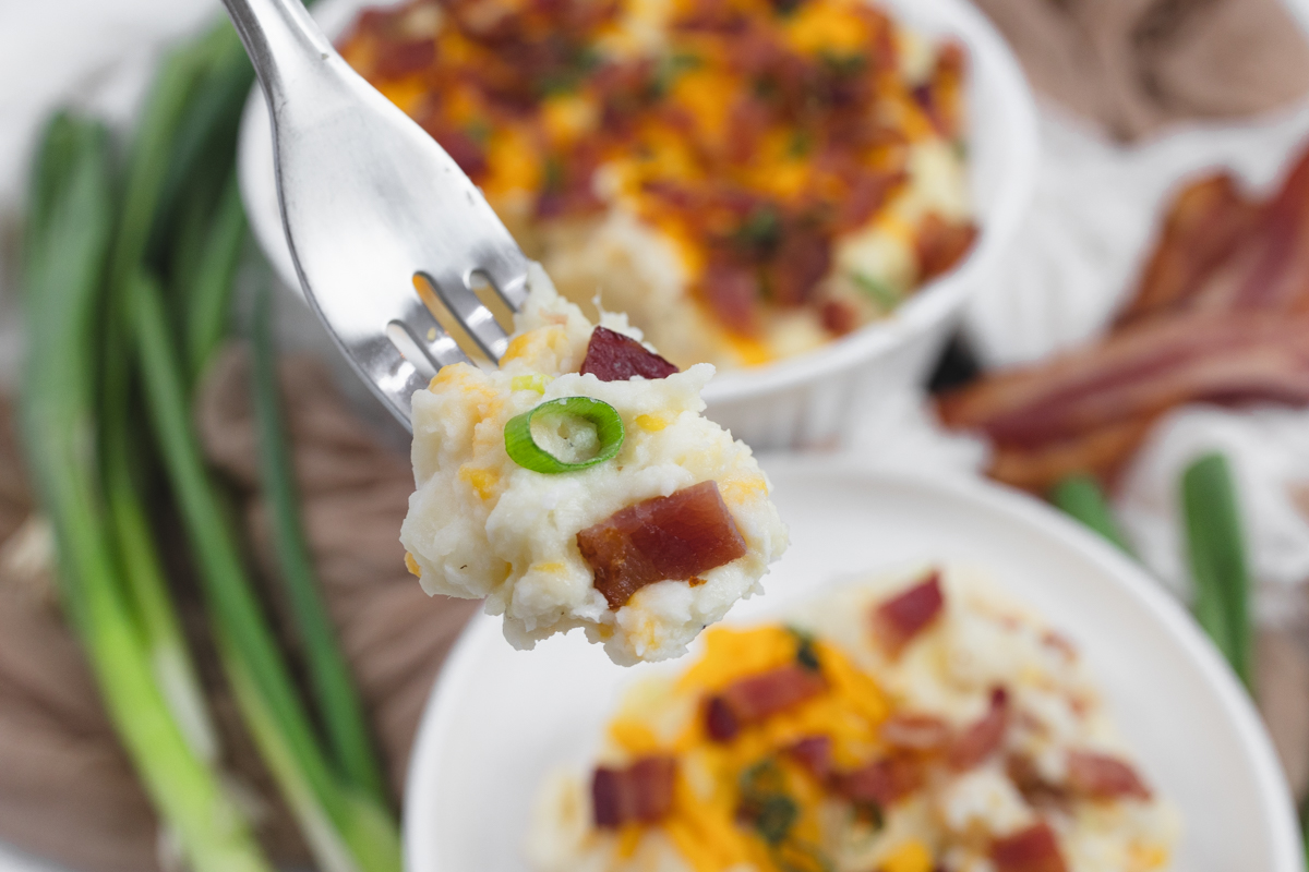 Close up shot of a fork lifting up a bite of loaded mashed potatoes, with the plate in the background.