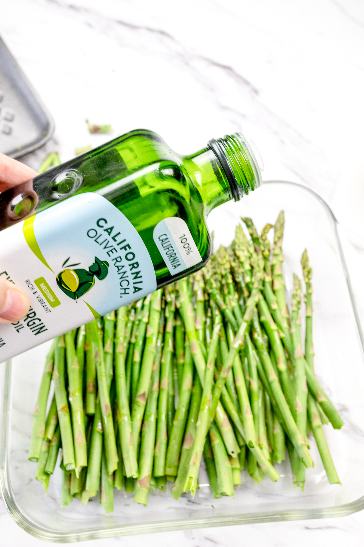Close up shot of olive oil being drizzled on asparagus in a glass dish.