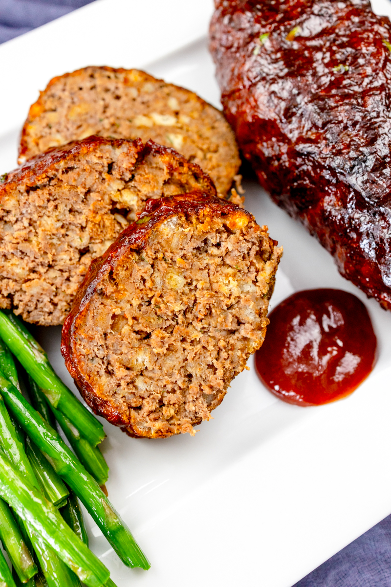 Close view of meatloaf slices on a white plate with asparagus and dipping sauce.