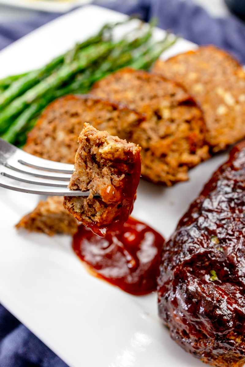 Close view of a fork holding a piece of meatloaf in front of meatloaf slices on a white plate with asparagus and dipping sauce.