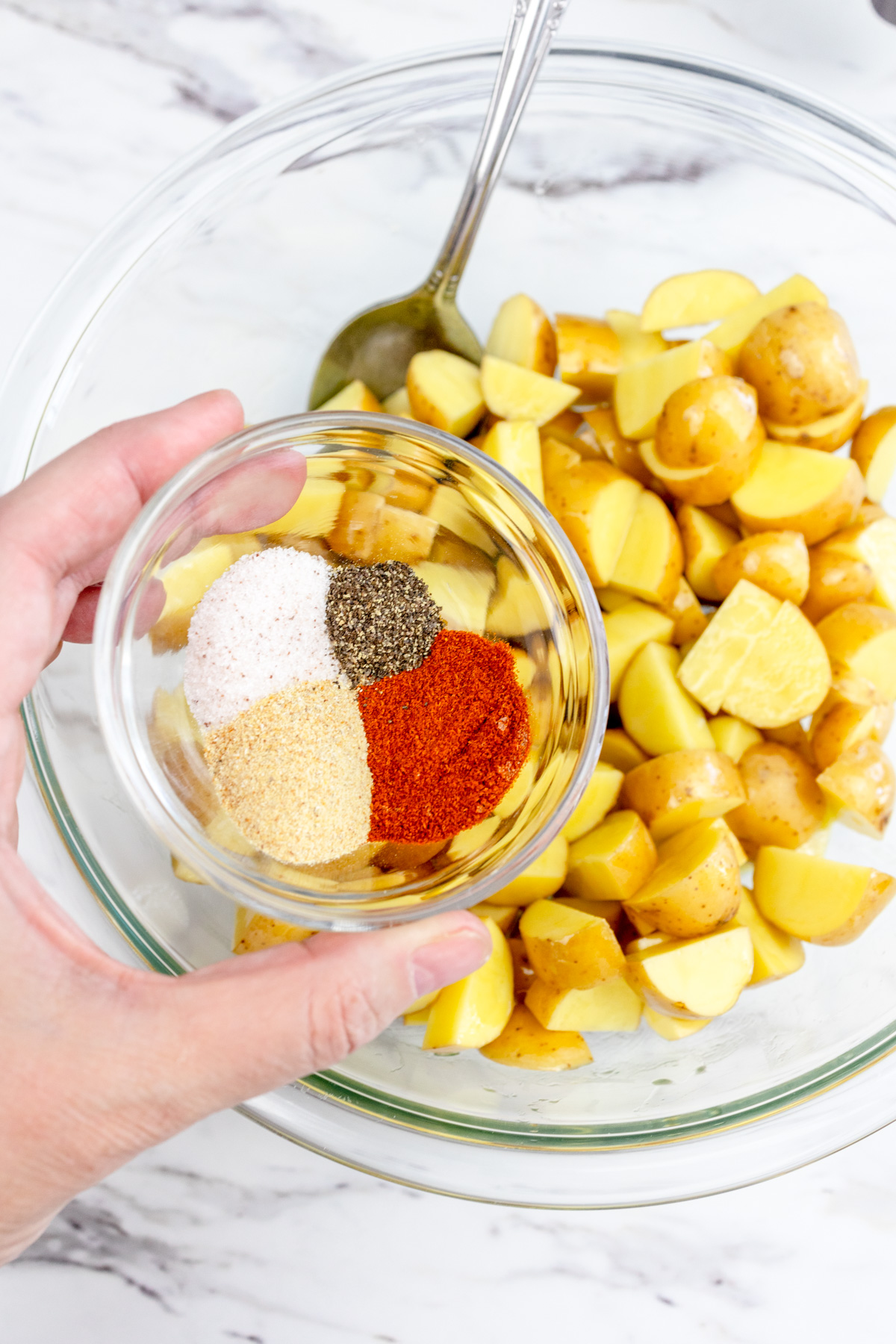 A hand holding the seasonings in a small bowl over the potatoes in the large mixing bowl, ready to mix together.