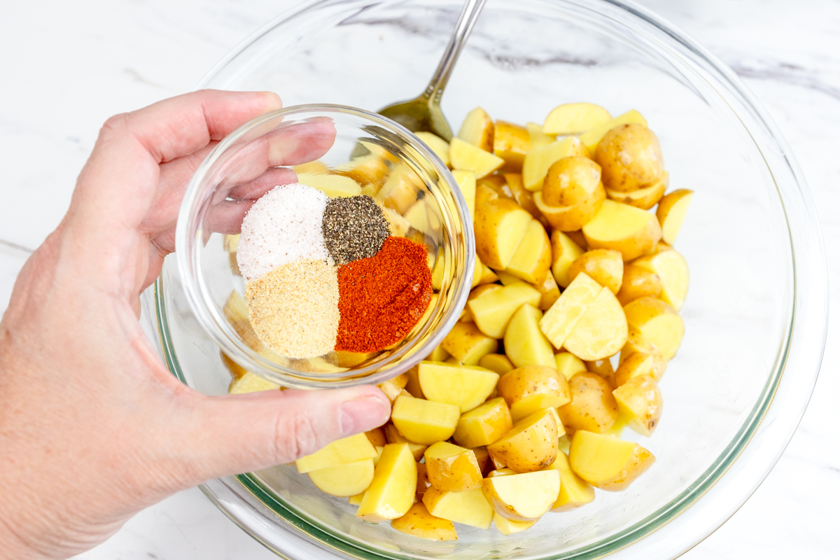 A hand holding the seasonings in a small bowl over the potatoes in the large mixing bowl, ready to mix together.