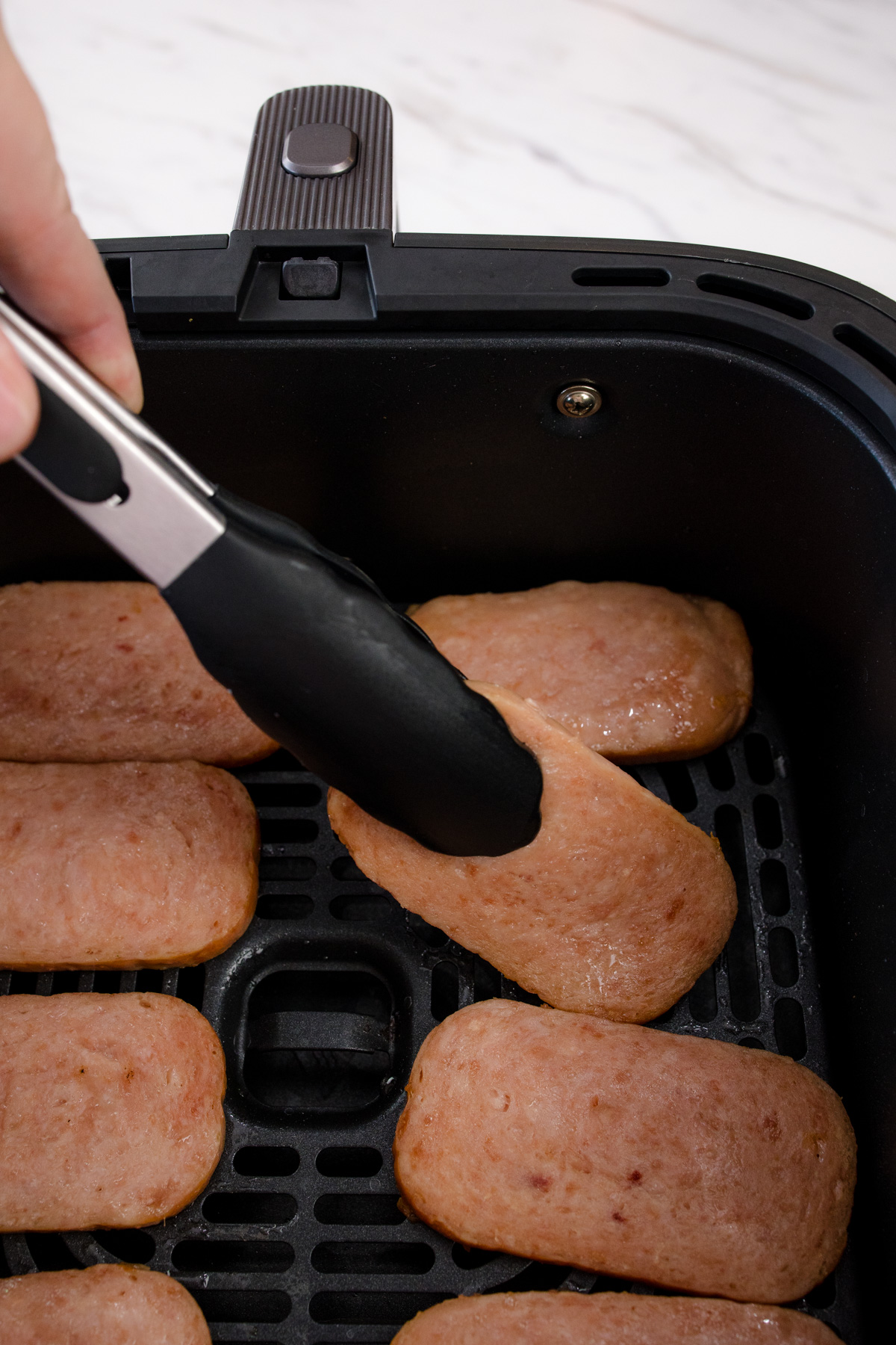 Tongs holding a slice of cooked spam in an air fryer basket amongst other spam slices in the basket.