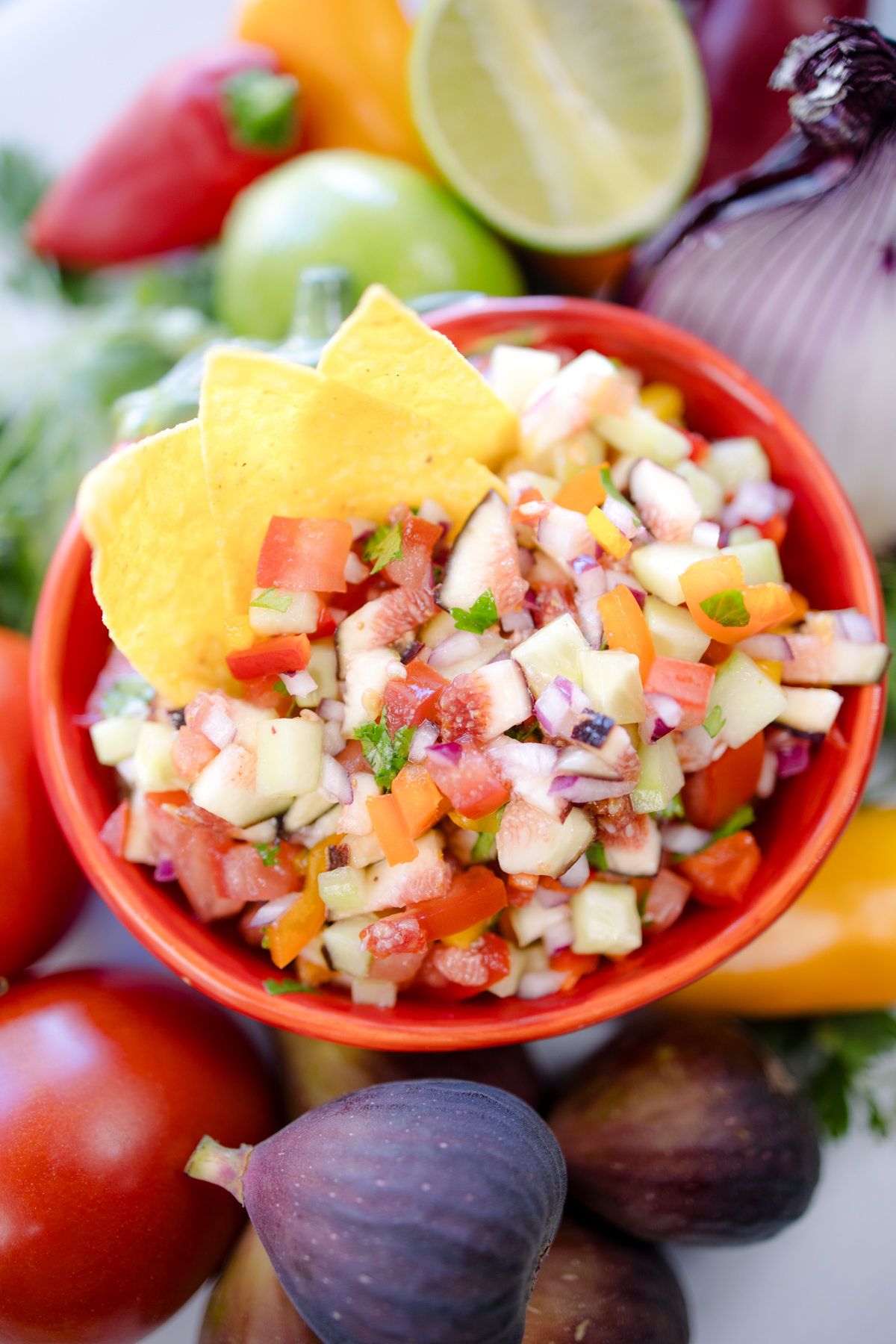 A bowl of Fig Salsa surrounded by fresh ingredients, with tortilla chips inserted into the salsa.