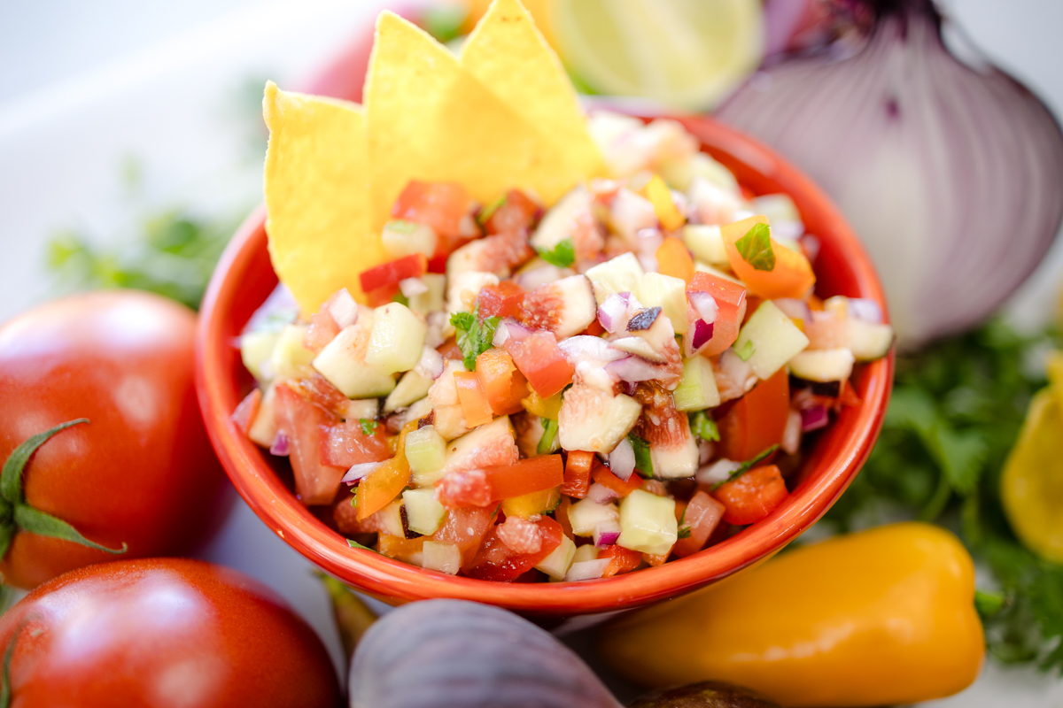 A bowl of Fig Salsa surrounded by fresh ingredients, with tortilla chips inserted into the salsa.
