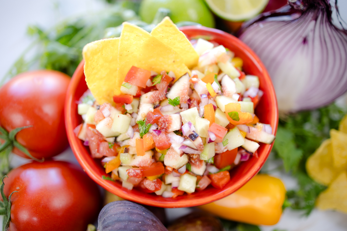 A bowl of Fig Salsa surrounded by fresh ingredients, with tortilla chips inserted into the salsa.