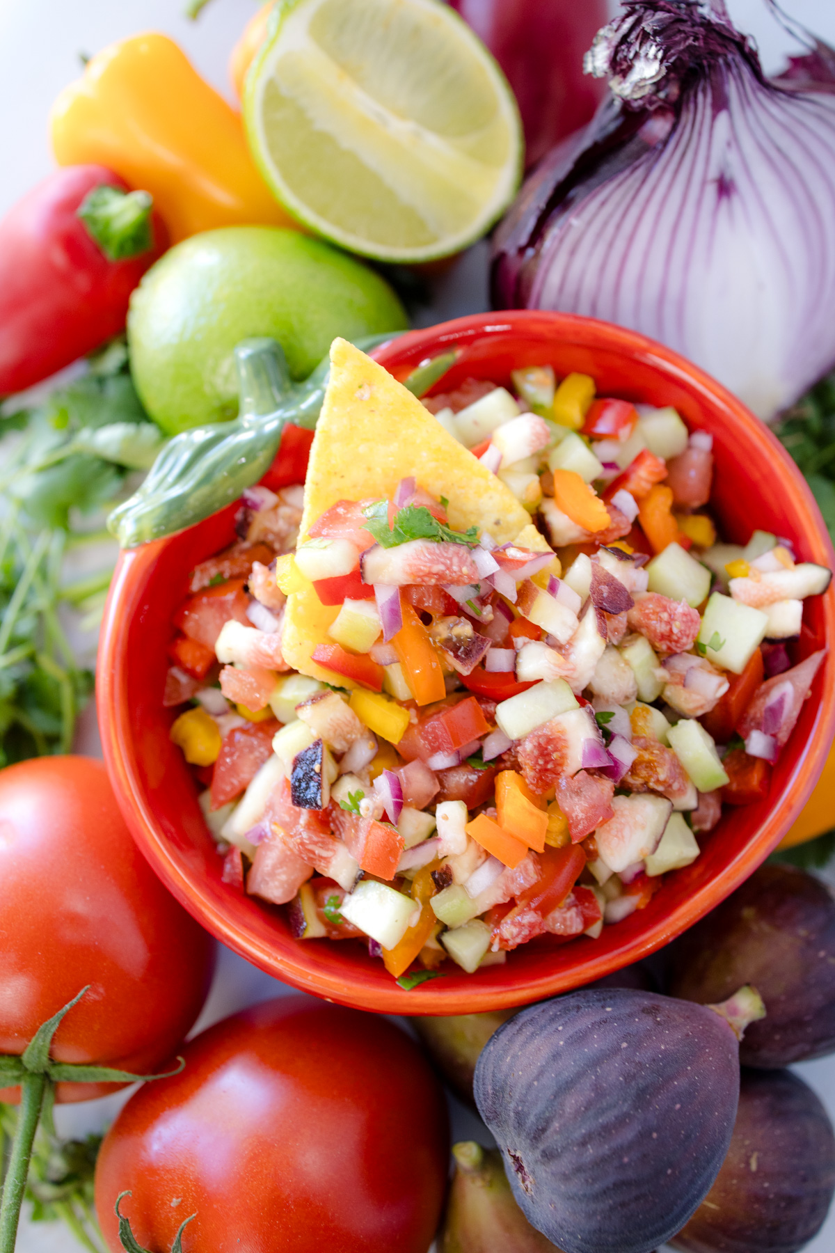 A bowl of Fig Salsa surrounded by fresh ingredients, with a tortilla chip inserted into the salsa.