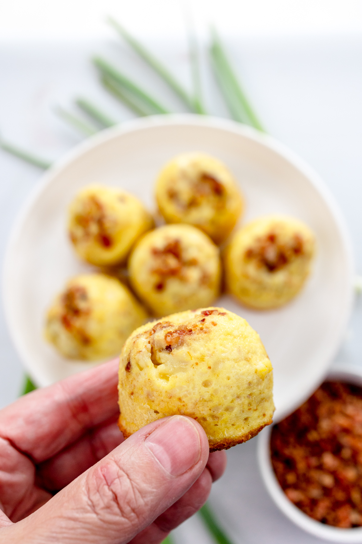 Close up shot of a hand holding an egg bite in front of Air Fryer Egg Bites on a white plate.