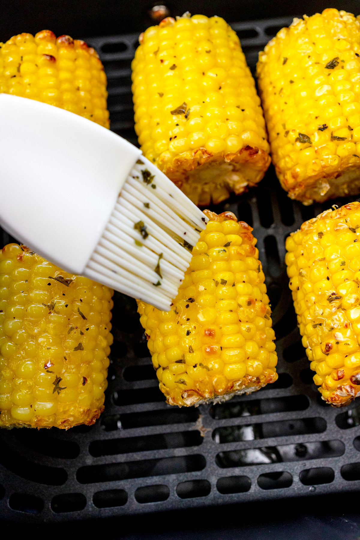 Close up of a pastry brush basting mini air fried corn on the cobs in an air fryer basket.