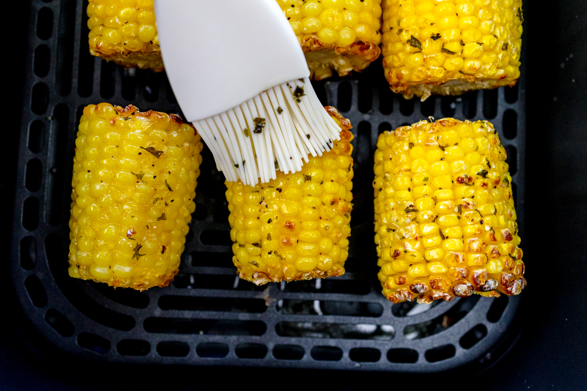 Close up of a pastry brush basting mini air fried corn on the cobs in an air fryer basket.