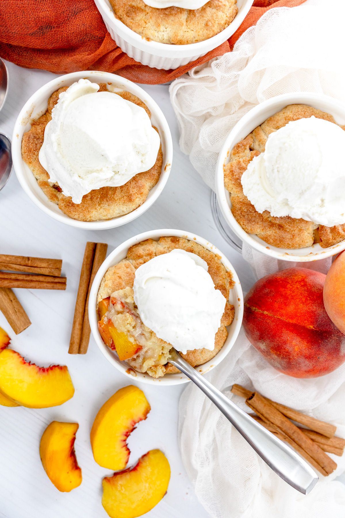 Overhead image of Air Fryer Peach Cobbler in ramekins with whipped cream on top, with peach slices and cinnamon sticks around them.