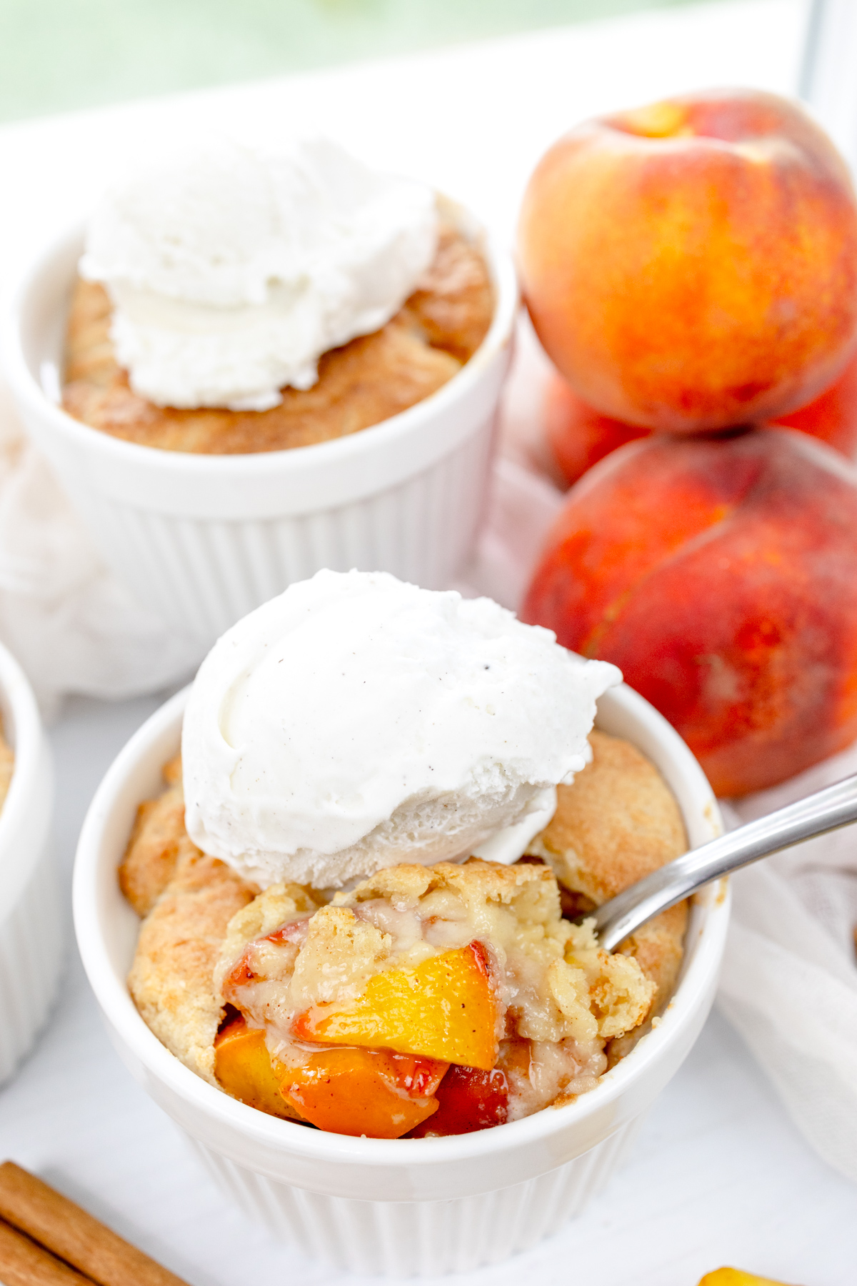 Angled image of Air Fryer Peach Cobbler in ramekins with whipped cream on top, with peaches and cinnamon sticks around them.