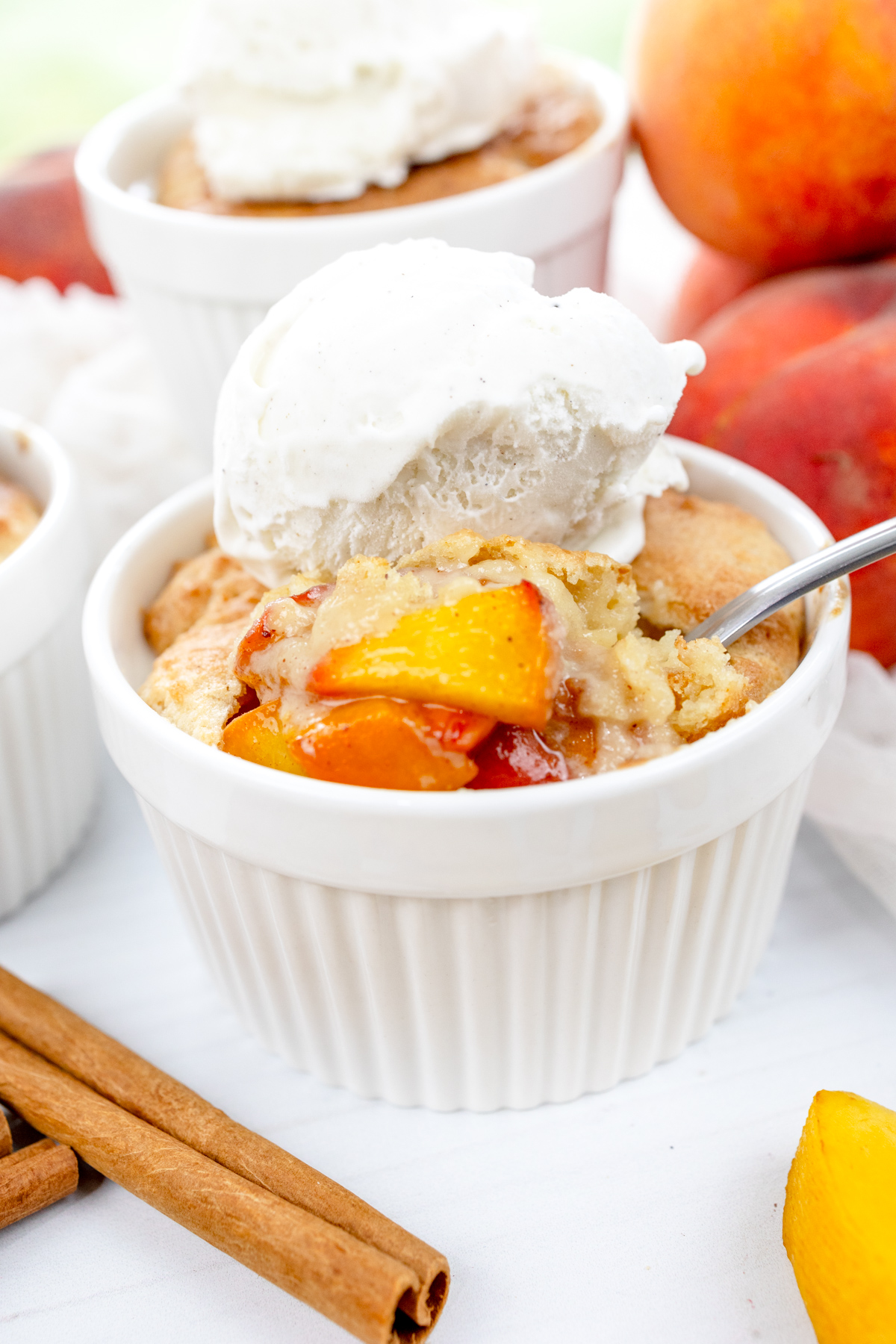 Angled image of Air Fryer Peach Cobbler in ramekins with whipped cream on top, with peach slices and cinnamon sticks around them.