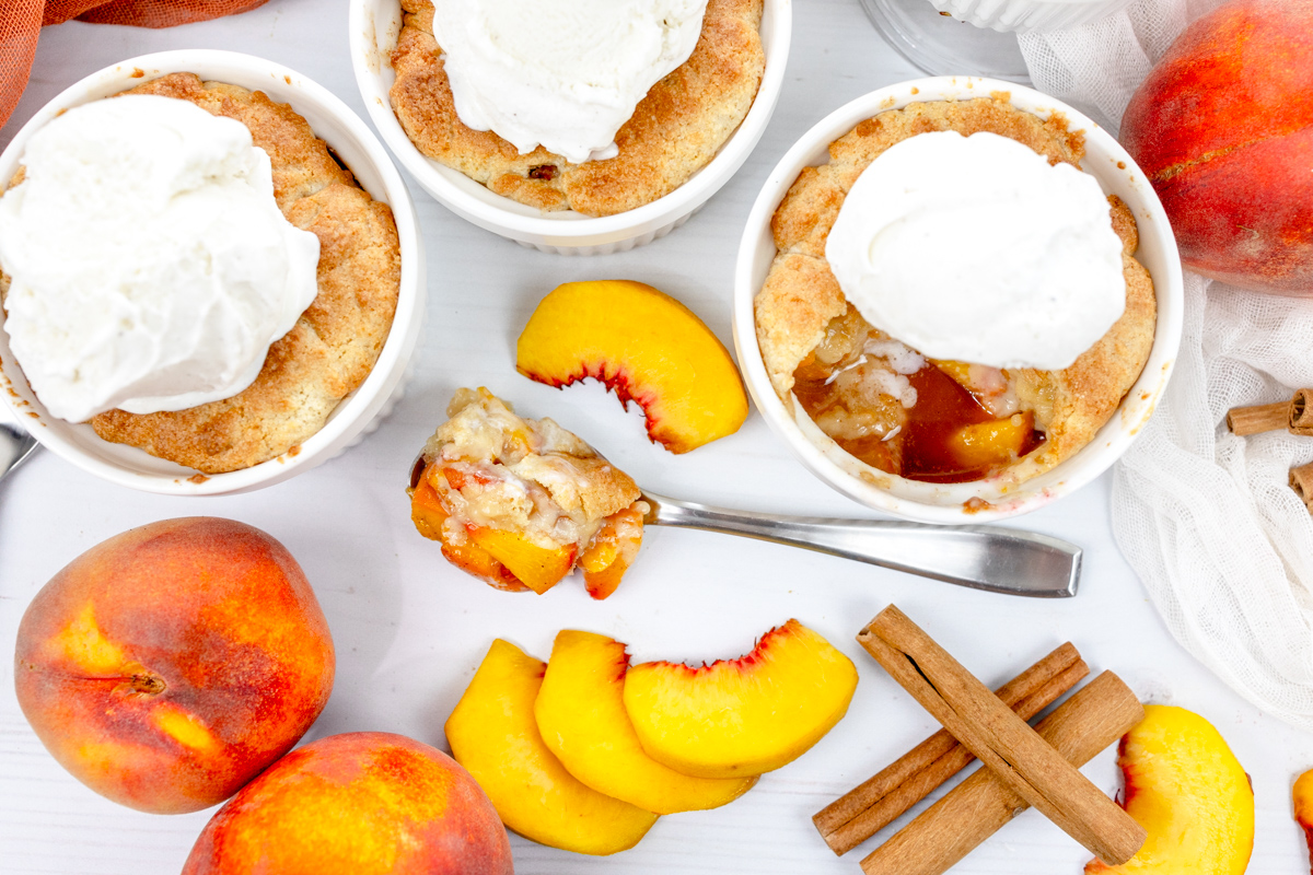 Overhead image of Air Fryer Peach Cobbler in ramekins with whipped cream on top, with peach slices and cinnamon sticks around them.