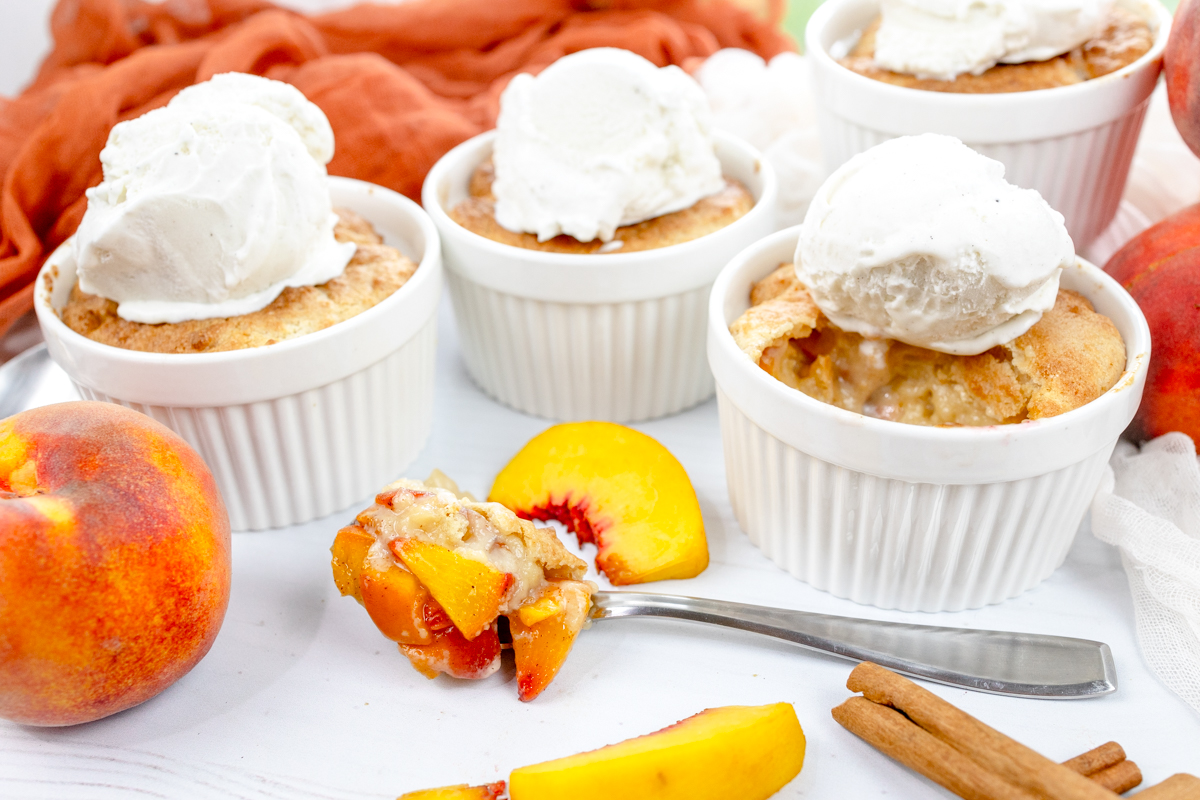 Angled image of Air Fryer Peach Cobbler in ramekins with whipped cream on top, with peach slices and cinnamon sticks around them.