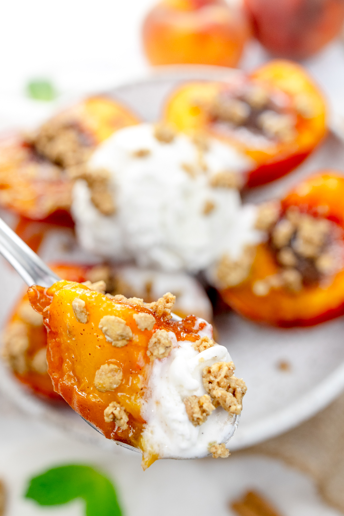 Close up image of Air Fryer Peaches on a white plate with a scoop of ice cream and crumb sprinkling on top, with a spoon in the foreground containing a bite of the dessert.