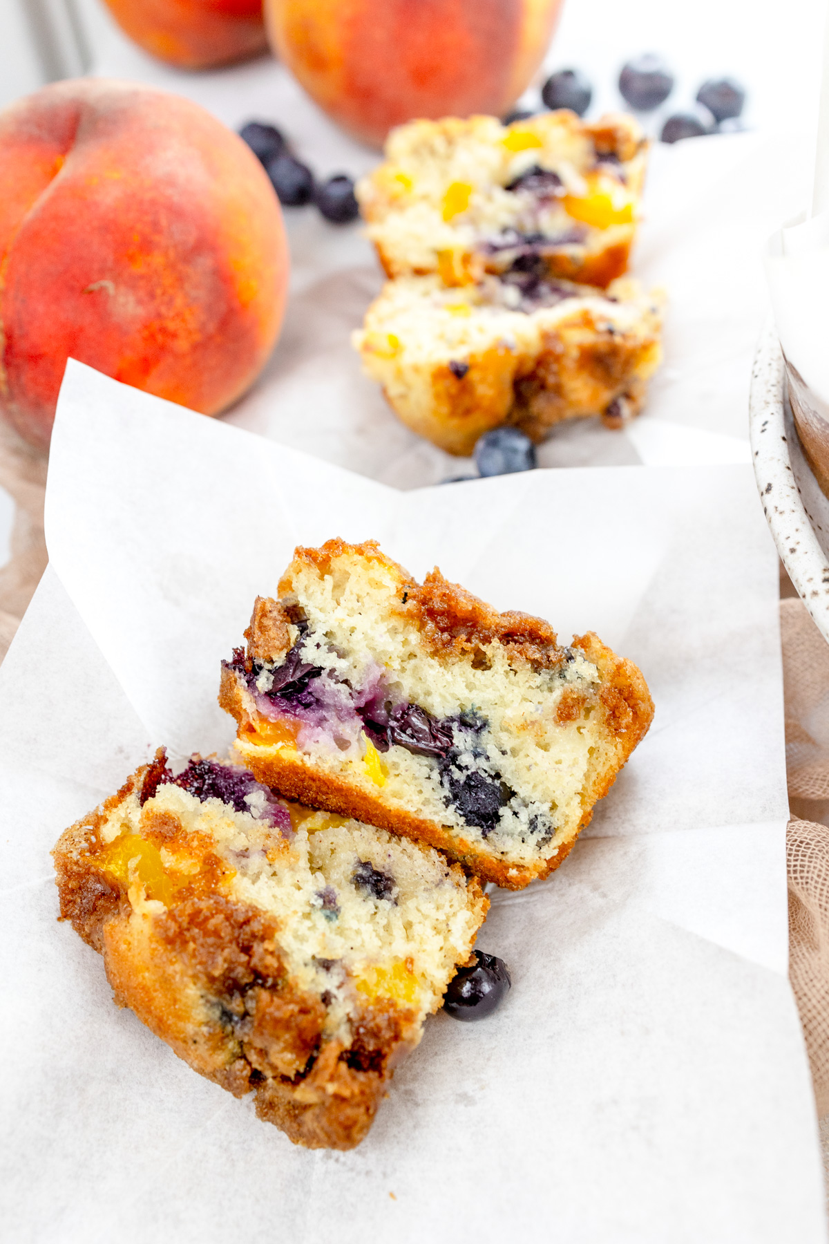 Overhead image of a Blueberry Peach Muffin that's been sliced in half, on a folded out paper muffin case, next to peaches, blueberries, and another muffin.