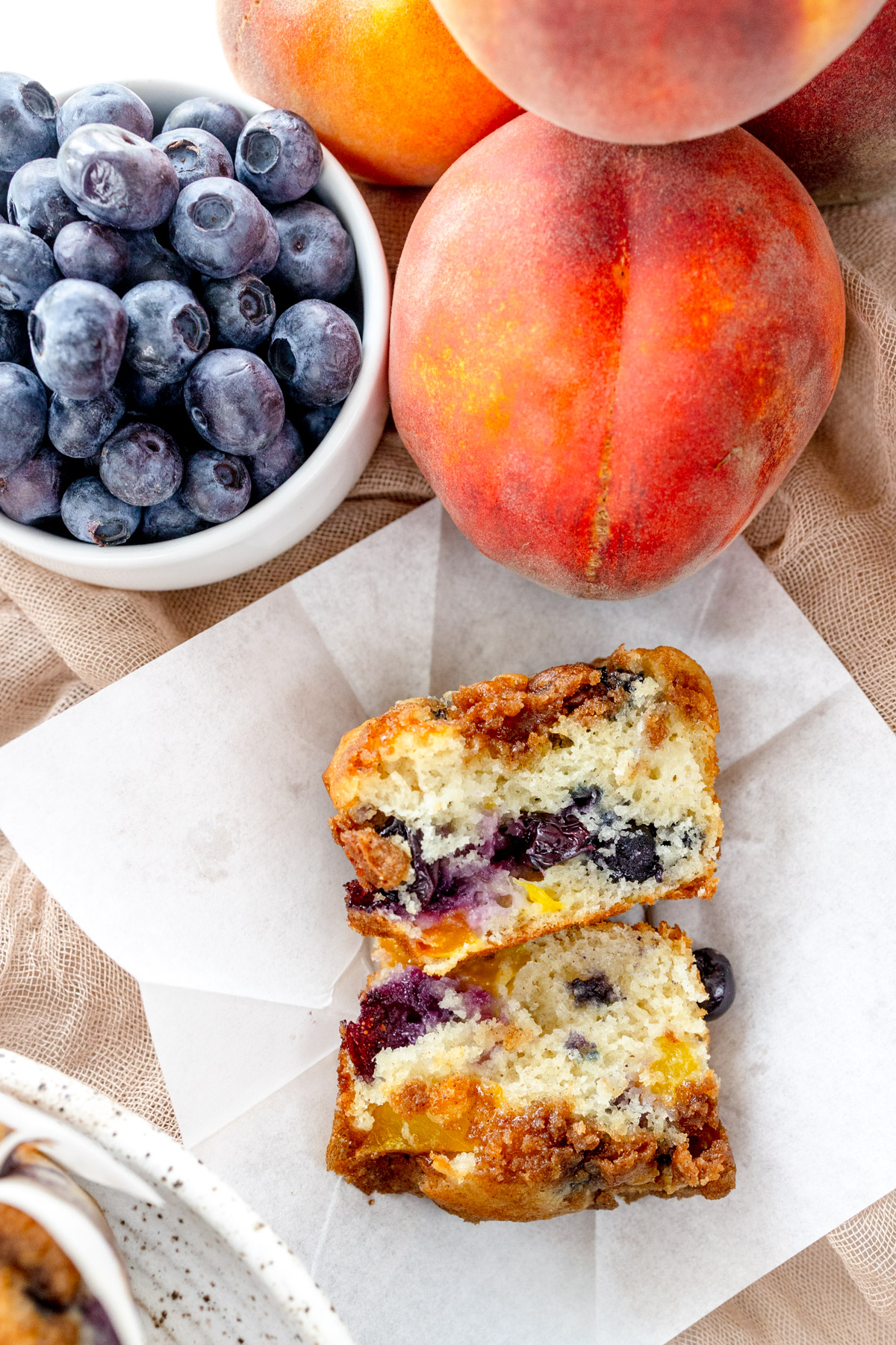 Overhead image of a Blueberry Peach Muffin that's been sliced in half, on a folded out paper muffin case, next to a peach and a bowl of blueberries.