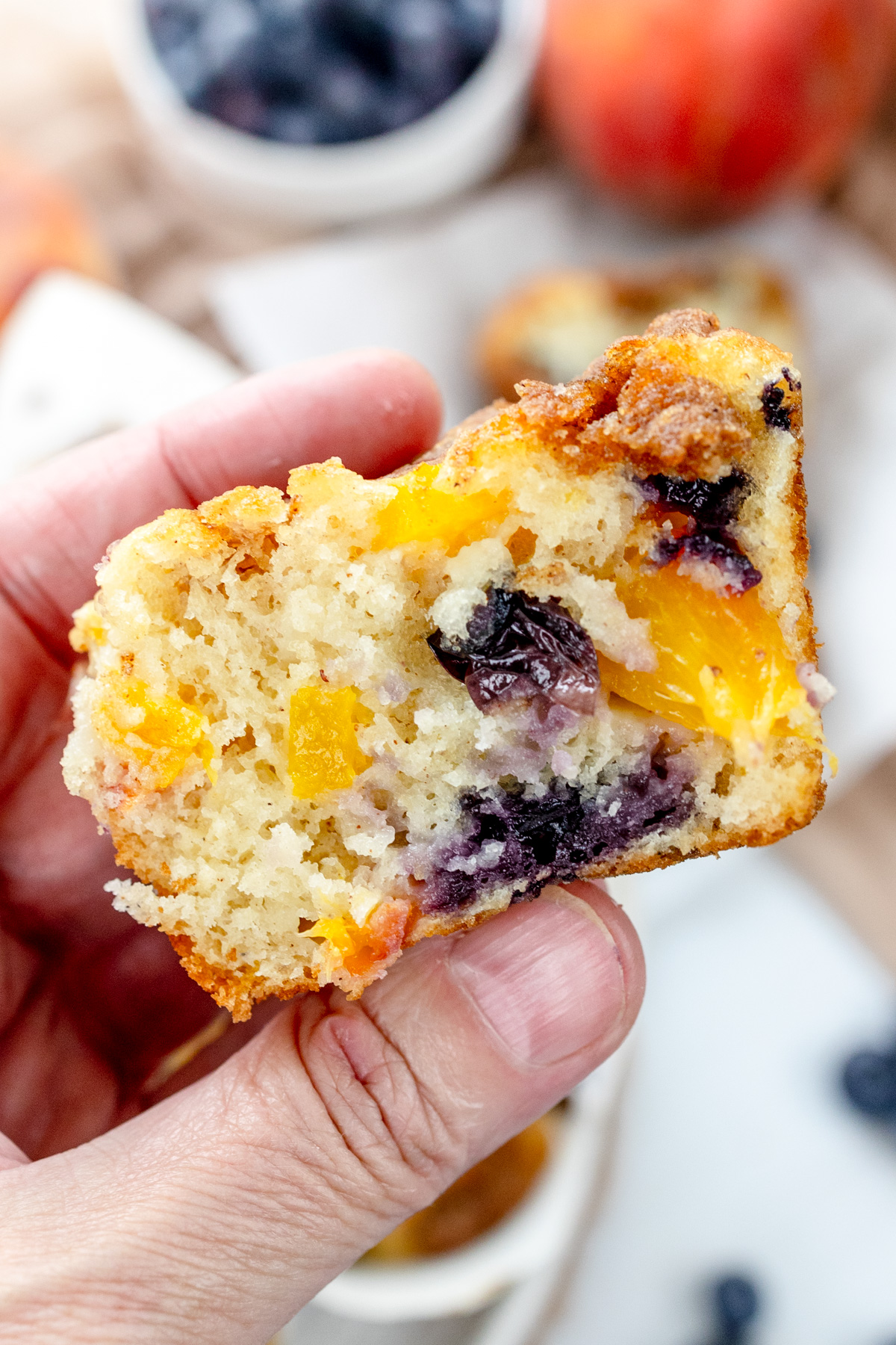 Close up image of a hand holding a Blueberry Peach Muffin that has been cut in half.