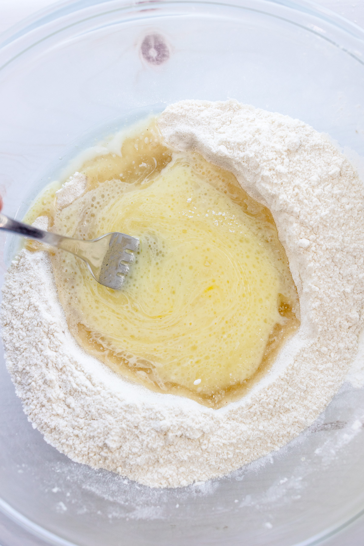 Close up image of a mixing bowl with dry ingredients needed for Cardamom Muffins, with wet ingredients on top being mixed by a fork.