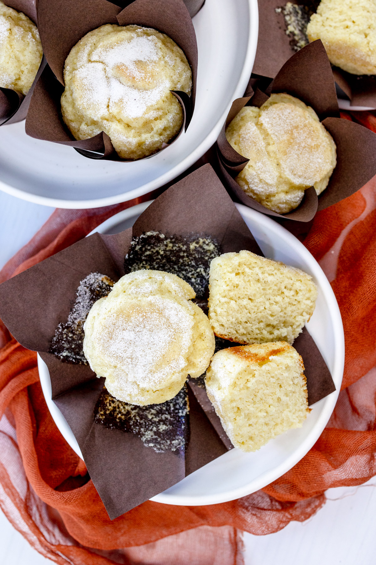 Overhead image of a Cardamom Muffin sliced in half, on an unfolded, brown paper muffin casing, next to another muffin, all on a white plate with other plates of muffins around it.