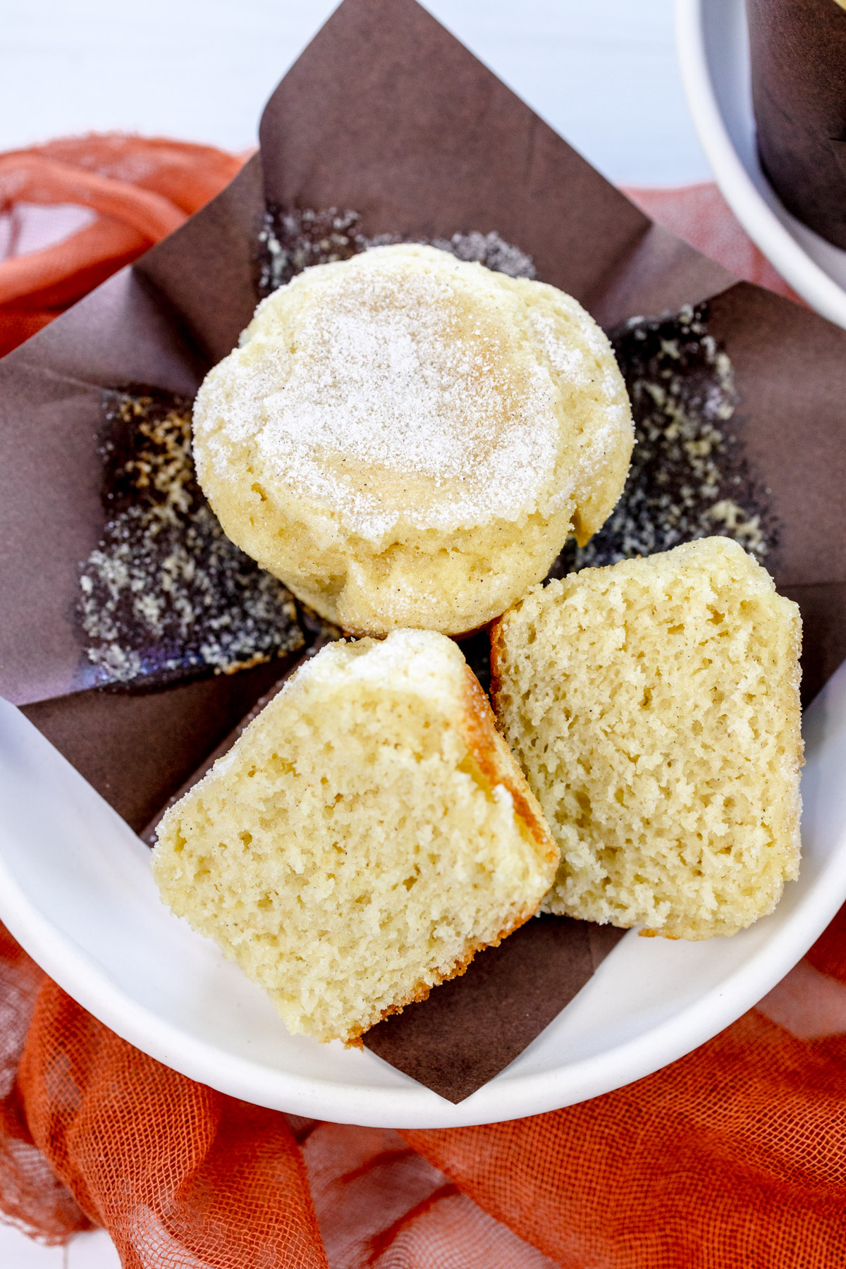 Close up image of a Cardamom Muffin sliced in half, on an unfolded, brown paper muffin casing, next to another muffin, all on a white plate.