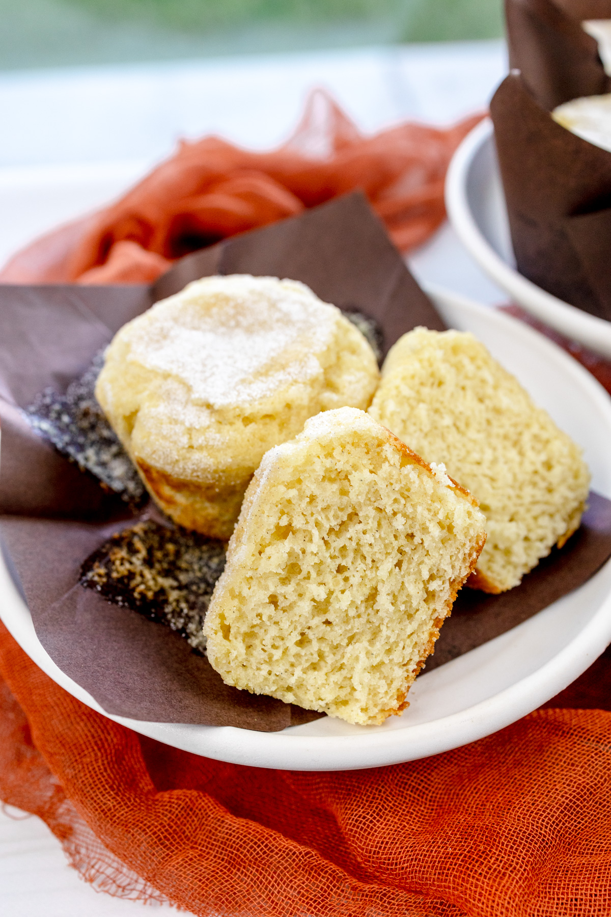 Close up image of a Cardamom Muffin sliced in half, on an unfolded, brown paper muffin casing, next to another muffin, all on a white plate.
