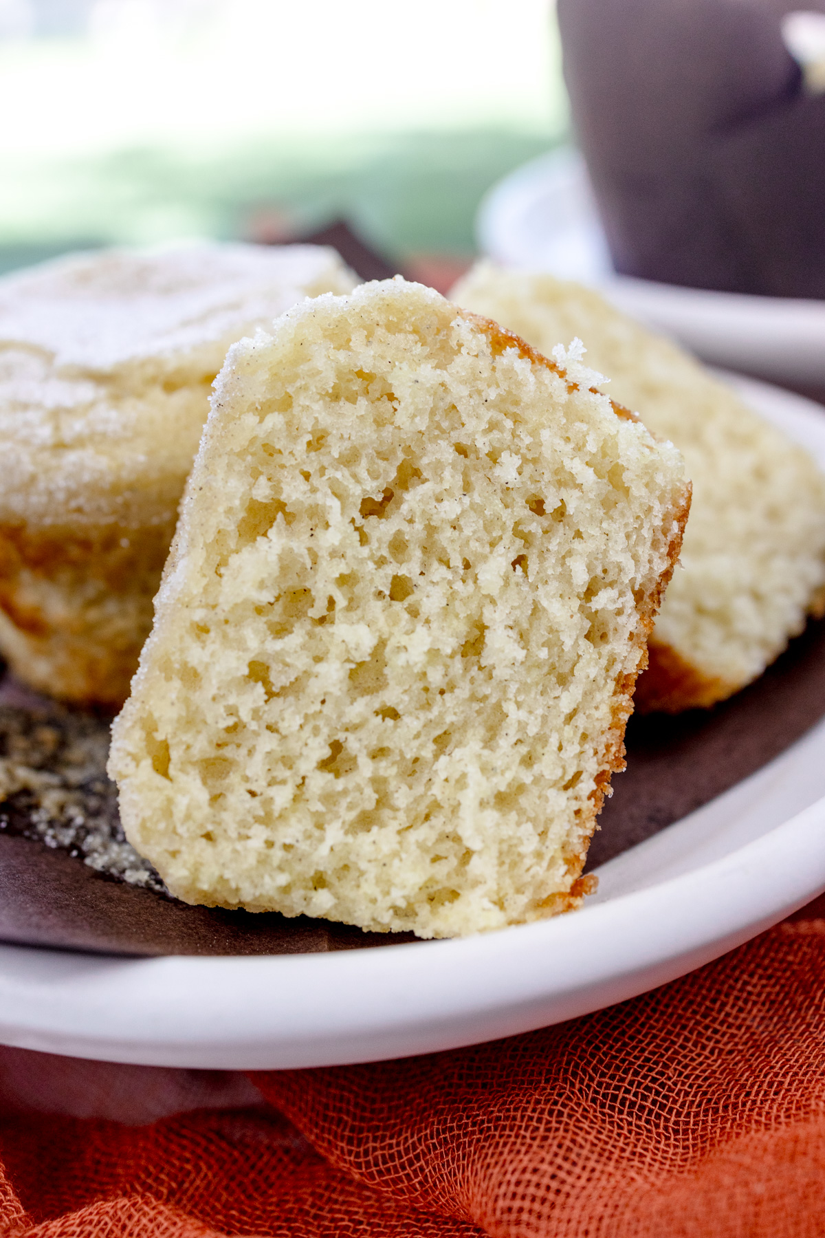 Close up image of a Cardamom Muffin sliced in half, on an unfolded, brown paper muffin casing, next to another muffin, all on a white plate.