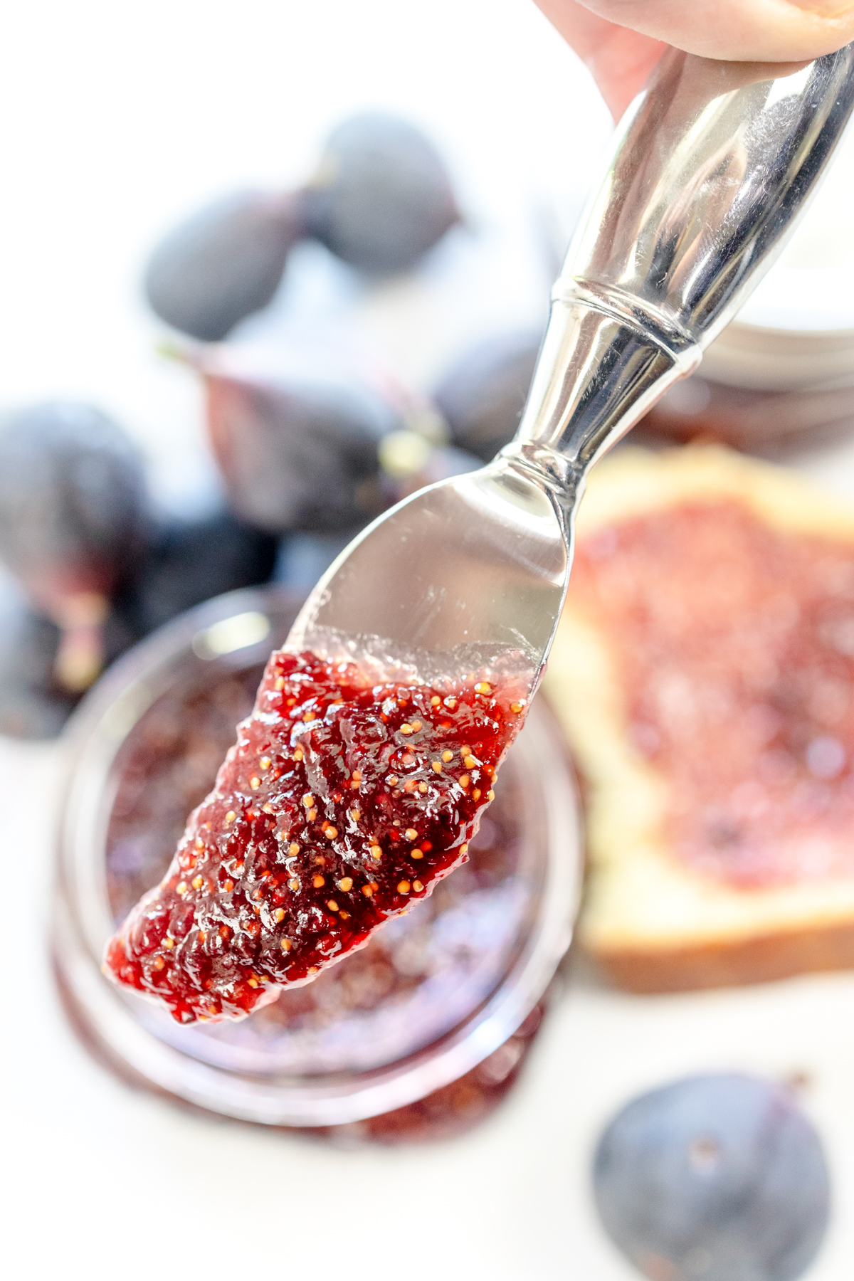A close up view of a spreading knife with fig jam on it, in front of a jar of fig jam and a piece of white bread with fig jam on it.