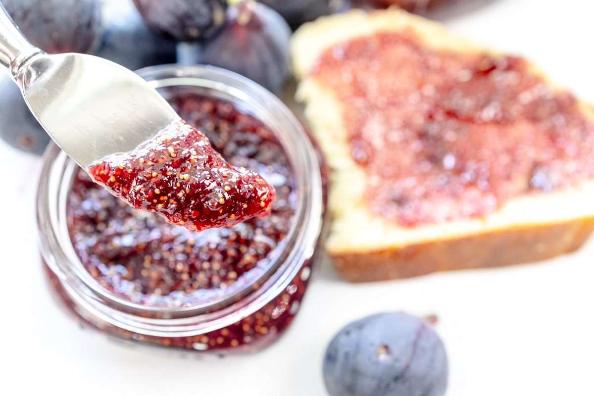 A close up view of a spreading knife with fig jam on it, in front of a jar of fig jam and a piece of white bread with fig jam on it.