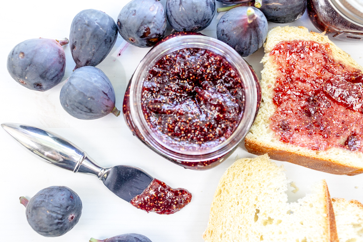 A close up view of a spreading knife with fig jam on it, next to a jar of fig jam and a piece of white bread with fig jam on it.