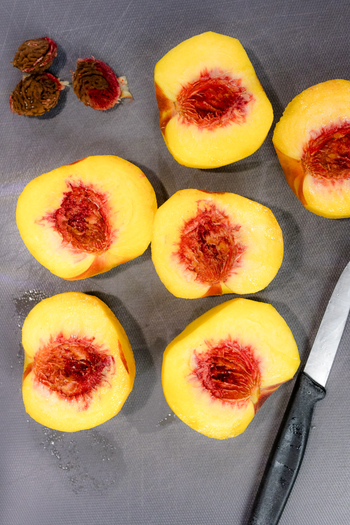Overhead view of sliced and pitted peaches on a dark chopping board next to a knife and the removed stones from the peaches.